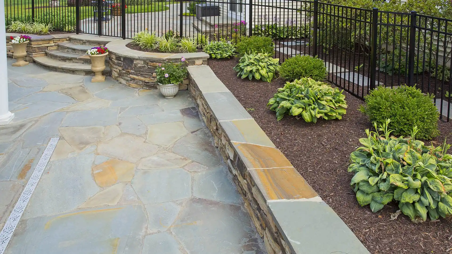 A landscaped garden with stone paving, a low stone wall, and stone steps leading up showcases the elegance of outdoor living in Delaware County, PA. Bordered by a black metal fence, lush green plants and flowers adorn the right side in soil beds.