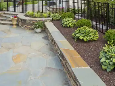 A landscaped garden with stone paving, a low stone wall, and stone steps leading up showcases the elegance of outdoor living in Delaware County, PA. Bordered by a black metal fence, lush green plants and flowers adorn the right side in soil beds.