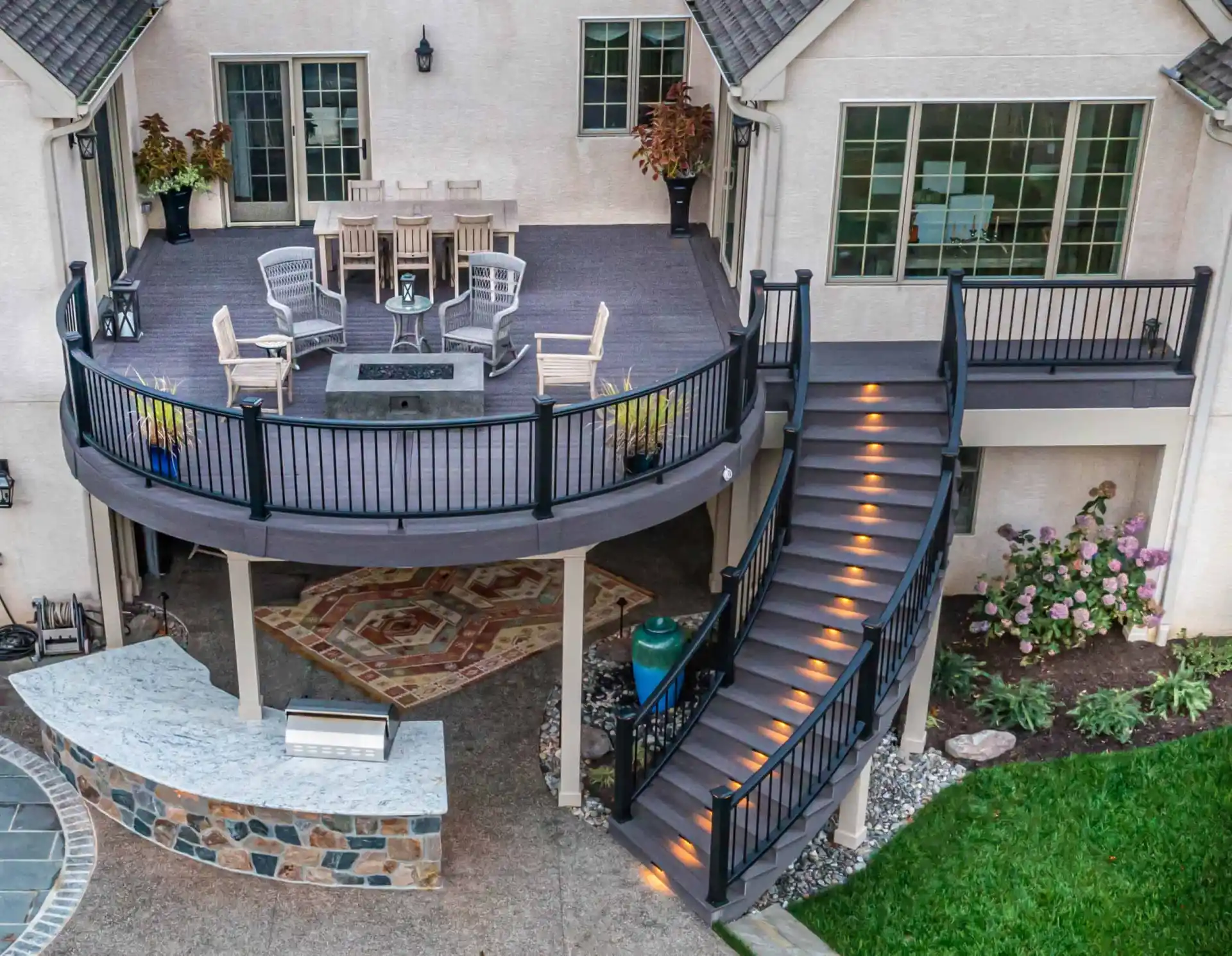 Aerial view of a two-story patio with a circular upper deck featuring a dining table, chairs, and fire pit. Curved staircase with glowing steps leads to the lower area with a bar and stone flooring, exemplifying premier outdoor living in Delaware County, PA. Vibrant green lawn in the foreground.