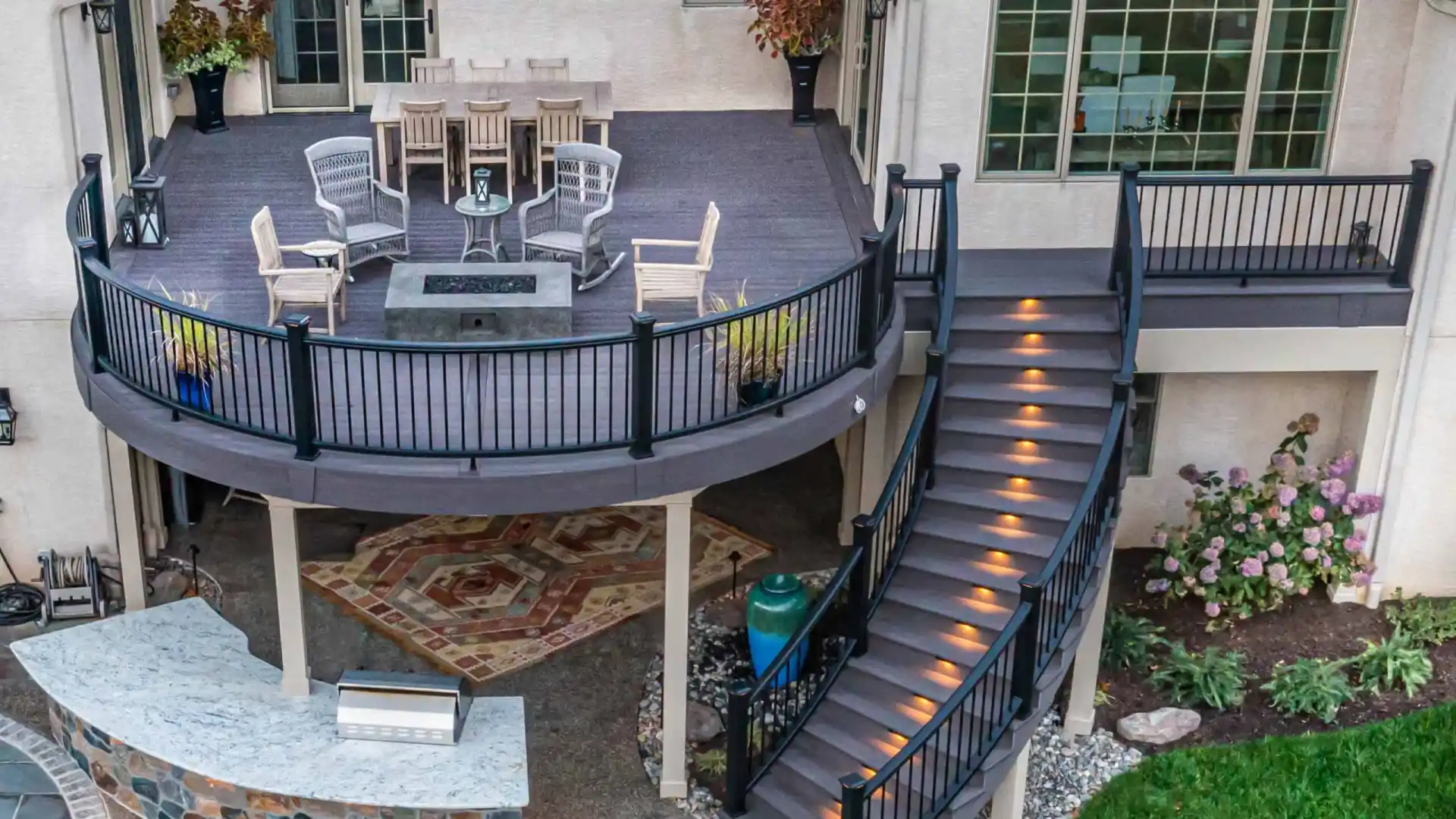 Aerial view of a two-story patio with a circular upper deck featuring a dining table, chairs, and fire pit. Curved staircase with glowing steps leads to the lower area with a bar and stone flooring, exemplifying premier outdoor living in Delaware County, PA. Vibrant green lawn in the foreground.