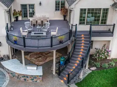 Aerial view of a two-story patio with a circular upper deck featuring a dining table, chairs, and fire pit. Curved staircase with glowing steps leads to the lower area with a bar and stone flooring, exemplifying premier outdoor living in Delaware County, PA. Vibrant green lawn in the foreground.
