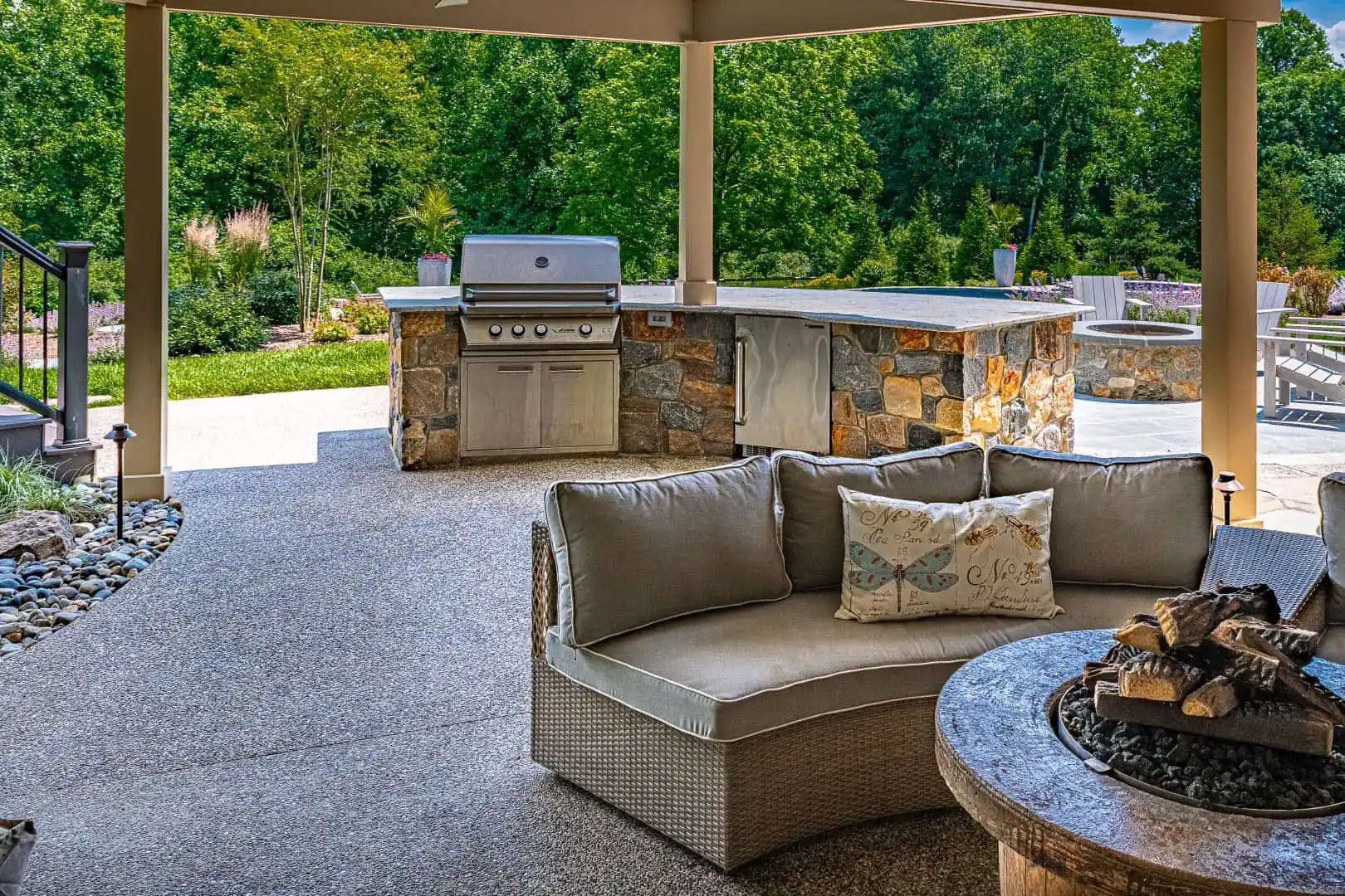 This inviting outdoor patio features a cushioned sofa encircling a stone fire pit, perfect for relaxing. In the backdrop, you'll find a stone barbecue grill and countertop set amidst lush greenery, showcasing the meticulous touch of landscaping services in Delaware County, PA.