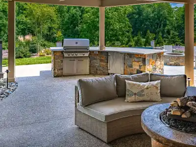 This inviting outdoor patio features a cushioned sofa encircling a stone fire pit, perfect for relaxing. In the backdrop, you'll find a stone barbecue grill and countertop set amidst lush greenery, showcasing the meticulous touch of landscaping services in Delaware County, PA.