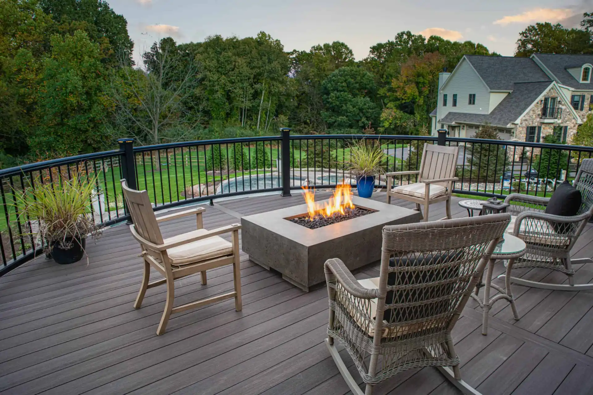 Four wicker chairs surround a lit rectangular fire pit on a spacious patio. With black railings and lush garden views, this Delaware County, PA haven showcases expert landscaping services. Potted plants accentuate the seating area, complementing the large house in the background.
