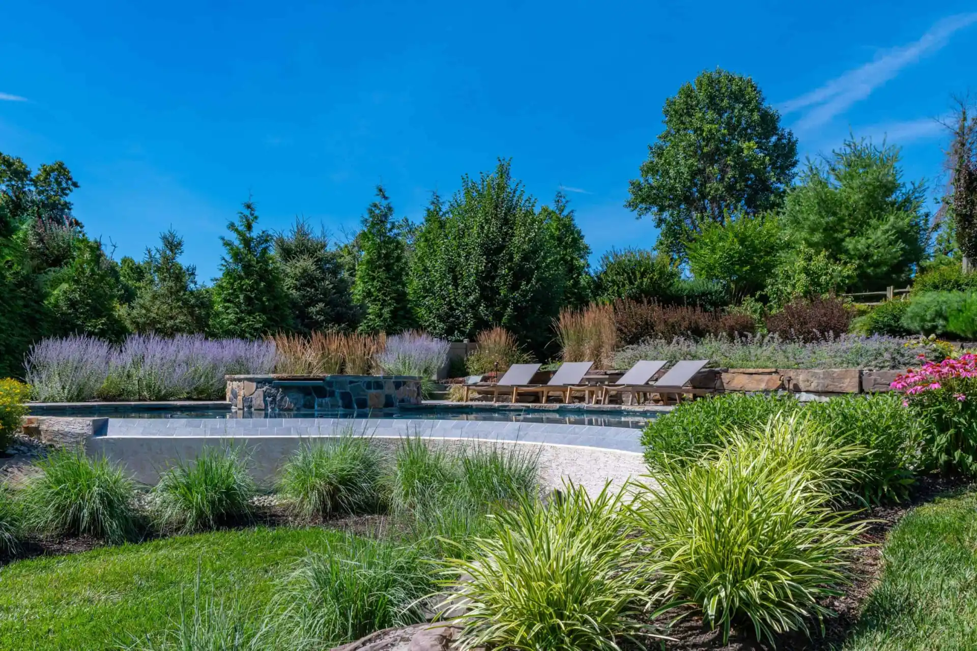A serene backyard in Delaware County, PA offers an enchanting outdoor living space with a stone-bordered pool surrounded by lush greenery and colorful plants. Several lounge chairs line the pool deck, and tall trees stand majestically under a clear blue sky, showcasing expert hardscaping services.