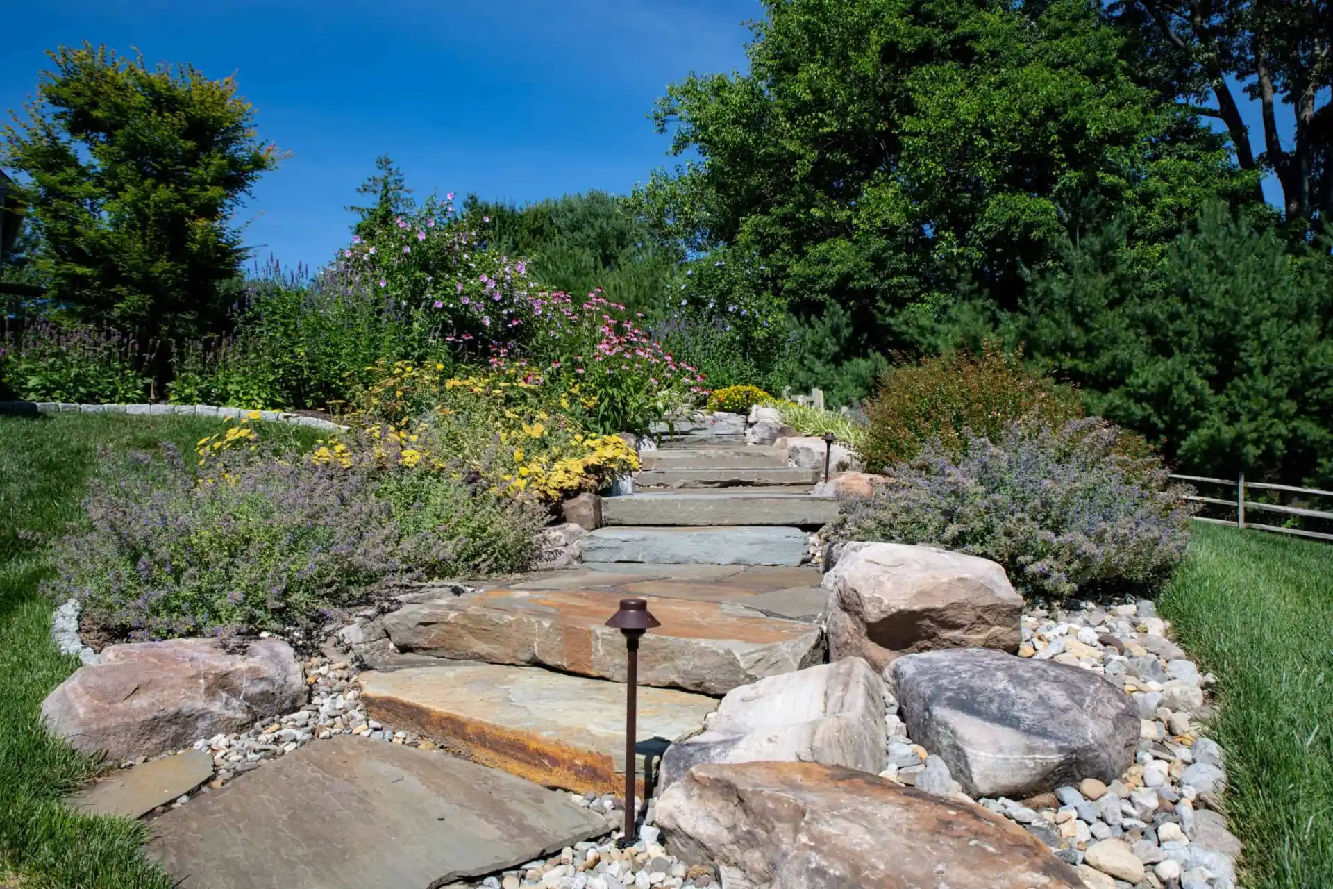 A stone path with large rocks leads uphill through a landscaped garden, flanked by colorful flowers and greenery. A single lamp is visible along the path. The sky is clear and blue, and trees border the area, showcasing the serene beauty of outdoor living in Delaware County, PA.
