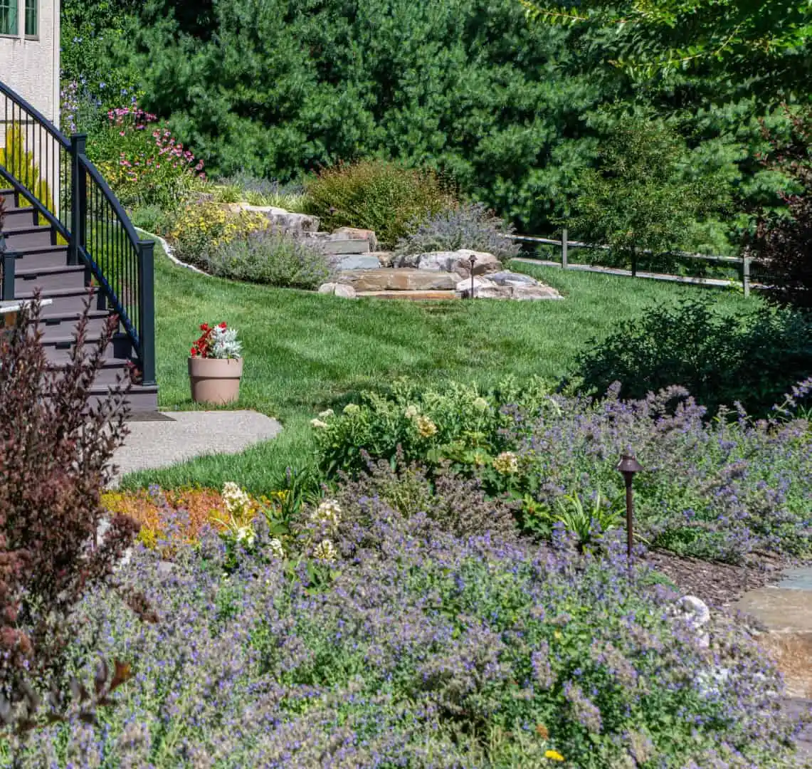 A landscaped garden with a stone path leading uphill, surrounded by blooming flowers and lush greenery. A pot of red flowers sits near the steps of a house on the left. With expert landscaping services in Delaware County, PA, enjoy trees and a wooden fence gracing the background.
