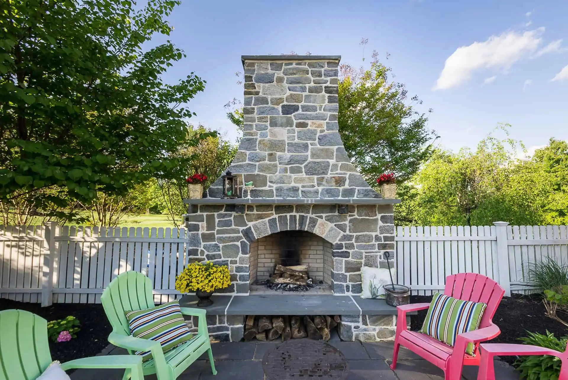 A stone outdoor fireplace with a woodpile below sits on a patio, surrounded by colorful Adirondack chairs and potted flowers, next to a white picket fence and lush green trees.