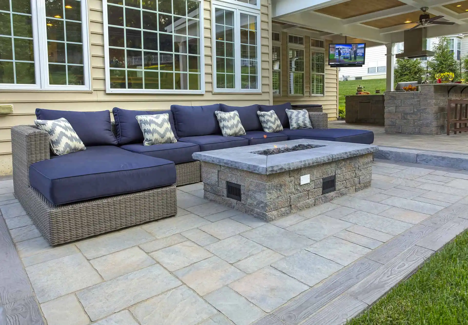 A spacious patio features a wicker sectional sofa with navy cushions and patterned pillows, centered around a stone fire pit, with an outdoor kitchen and TV in the background.