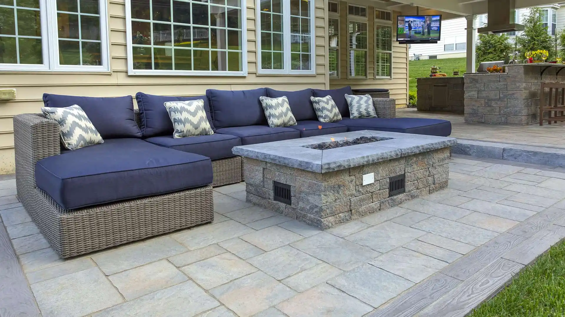Outdoor living in Delaware County, PA, comes to life with a patio showcasing a large wicker sectional with blue cushions and patterned pillows. A stone fire pit graces the paved surface, while a flat-screen TV and grill station complete this expertly designed oasis by hardscaping services.