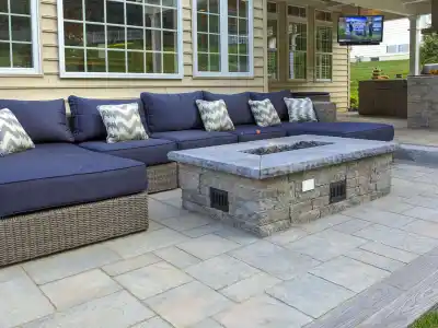 Outdoor living in Delaware County, PA, comes to life with a patio showcasing a large wicker sectional with blue cushions and patterned pillows. A stone fire pit graces the paved surface, while a flat-screen TV and grill station complete this expertly designed oasis by hardscaping services.