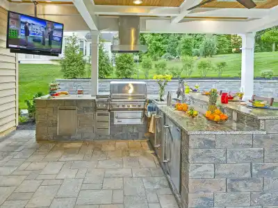 Outdoor living in Delaware County, PA offers a stunning kitchen equipped with a grill, oven, and granite countertop bar. A TV is mounted above the grill. Enjoy stone flooring surrounded by lush greenery and a retaining wall, perfect for hosting with fruits and drinks ready on the counter.