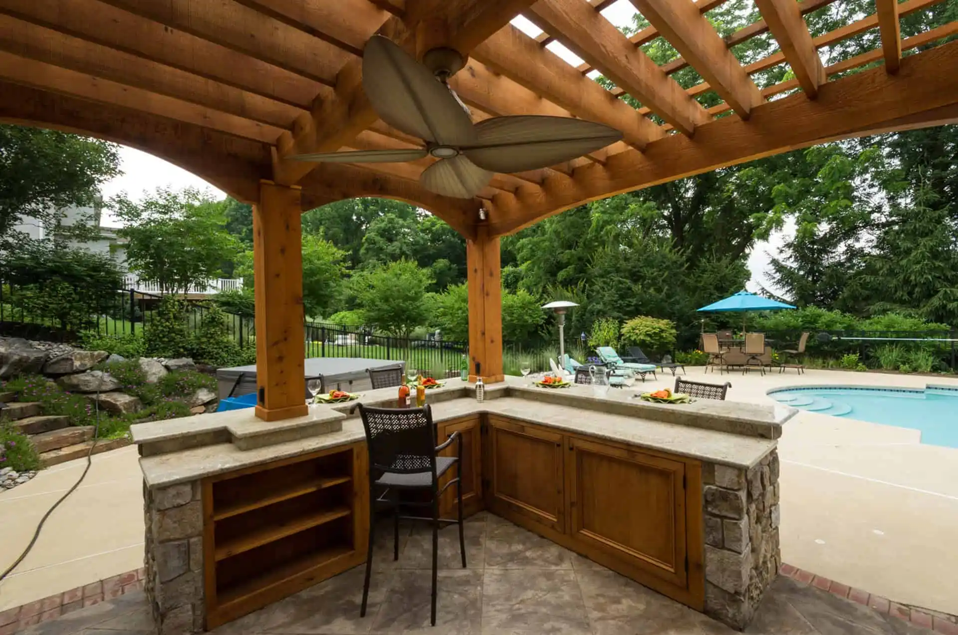 Outdoor kitchen with a granite countertop and wooden pergola overlooking a swimming pool, surrounded by greenery and lounge chairs with blue umbrellas. Two barstools sit at the counter.