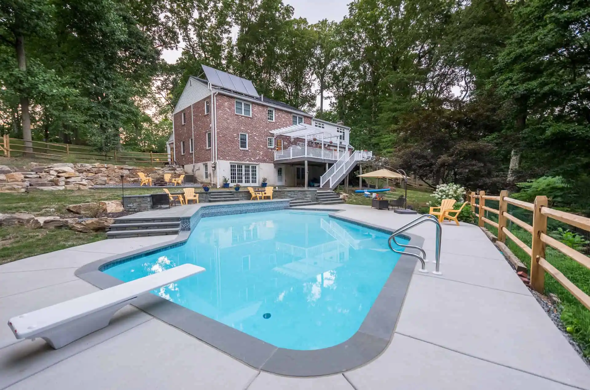 A backyard with a swimming pool, diving board, lounge chairs, umbrella, and a large brick house with solar panels, surrounded by trees and a wooden fence.