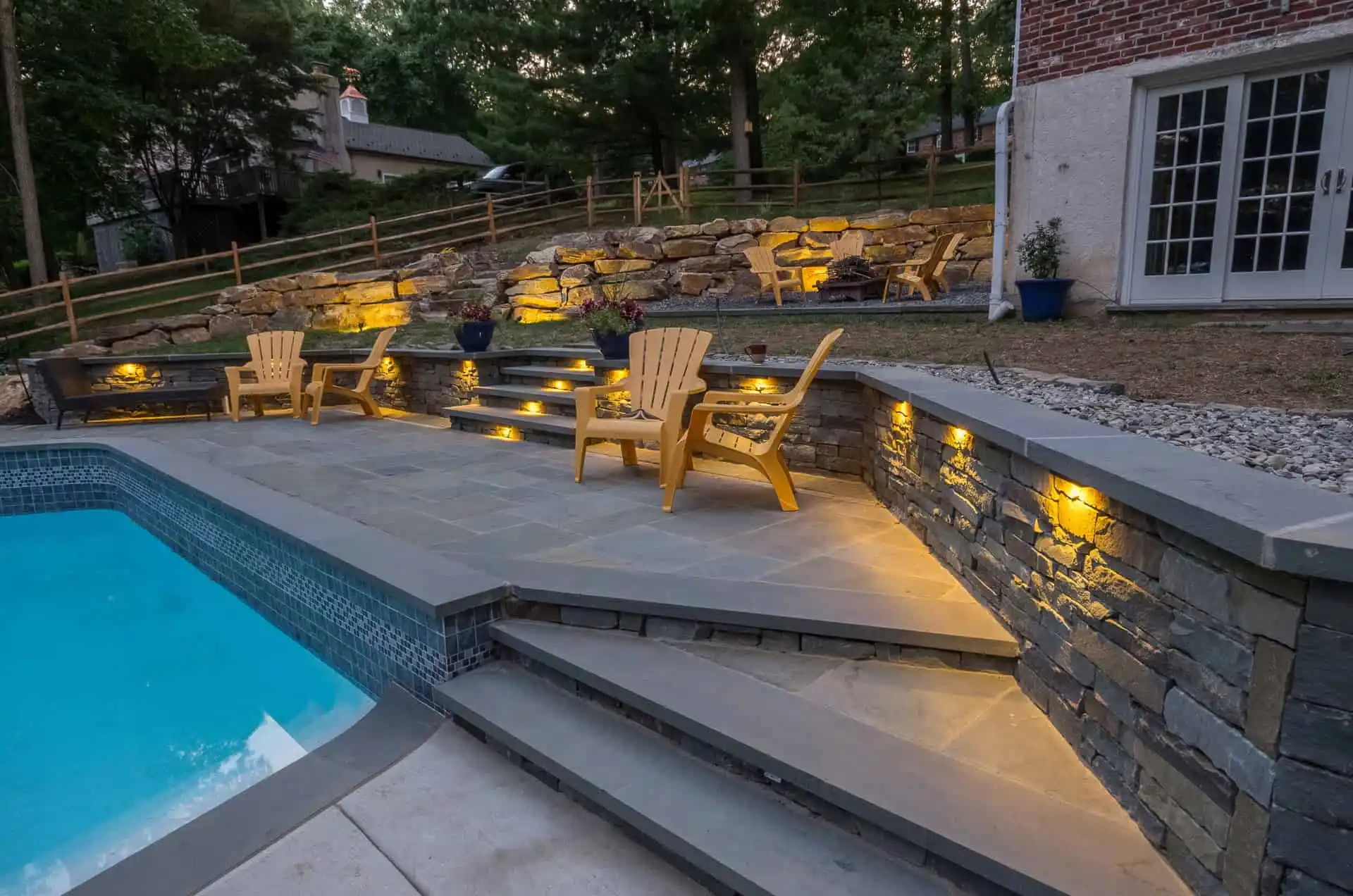 A backyard patio with stone steps and retaining walls, illuminated by built-in lights. Adirondack chairs are arranged near a swimming pool, with potted plants and trees in the background.
