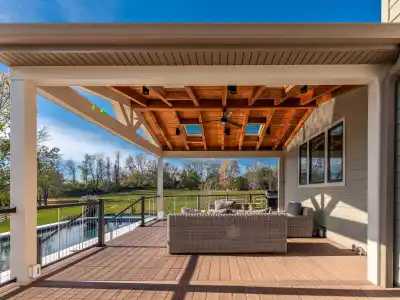 Experience outdoor living at its finest on a covered patio with a wooden ceiling and skylights. Wicker seating graces the wooden deck as you overlook the grassy yard and fenced-in pool beneath a clear blue sky. Perfect for those seeking exceptional landscaping services in Delaware County, PA.