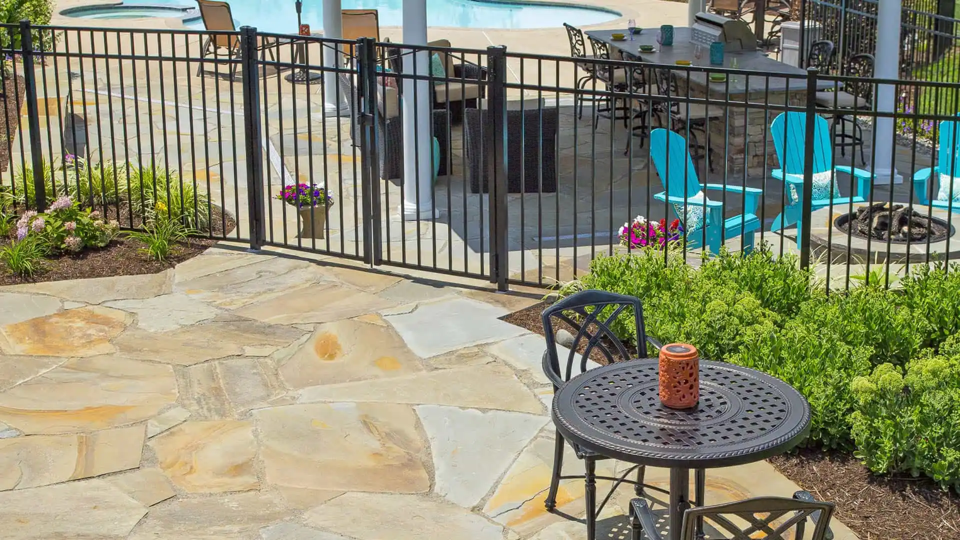 A patio featuring a round black metal table and chairs with a decorative orange candle, framed by expert landscaping services Delaware County PA. A black fence encloses the area, complete with a pool, blue seating, and stone fire pit. Green shrubs and vibrant flowers border the scene.