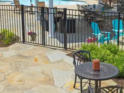 A patio featuring a round black metal table and chairs with a decorative orange candle, framed by expert landscaping services Delaware County PA. A black fence encloses the area, complete with a pool, blue seating, and stone fire pit. Green shrubs and vibrant flowers border the scene.