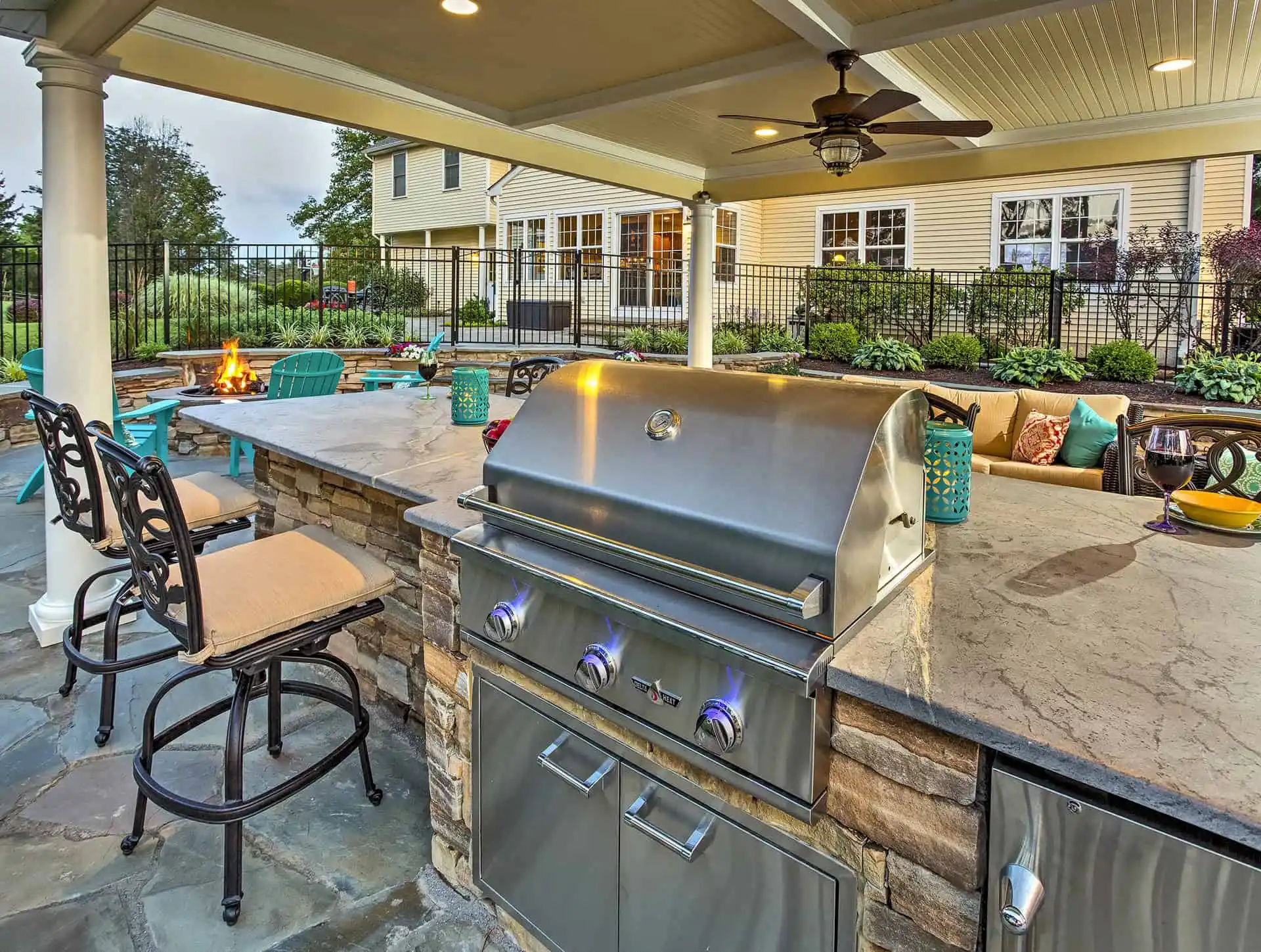 Covered outdoor kitchen and patio area featuring a stainless steel grill, stone countertops, bar seating, turquoise chairs, a fire pit, and a landscaped backyard with trees and shrubs.