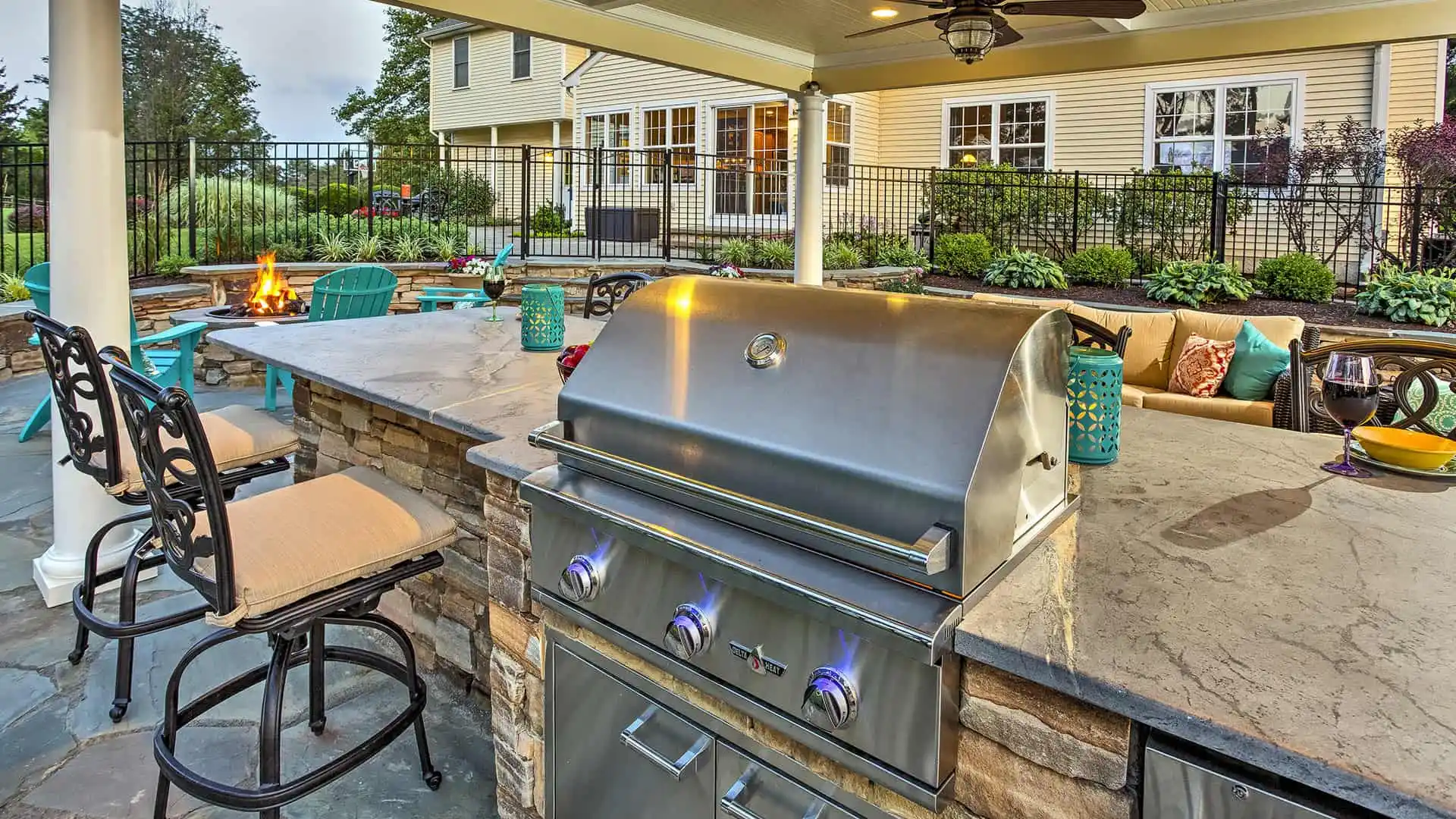 Outdoor living in Delaware County, PA, features a stunning outdoor kitchen with a stainless steel grill, stone countertops, and bar stools. A cozy seating area with a fire pit and decorative lanterns overlooks the garden and home behind a stylish black metal fence.