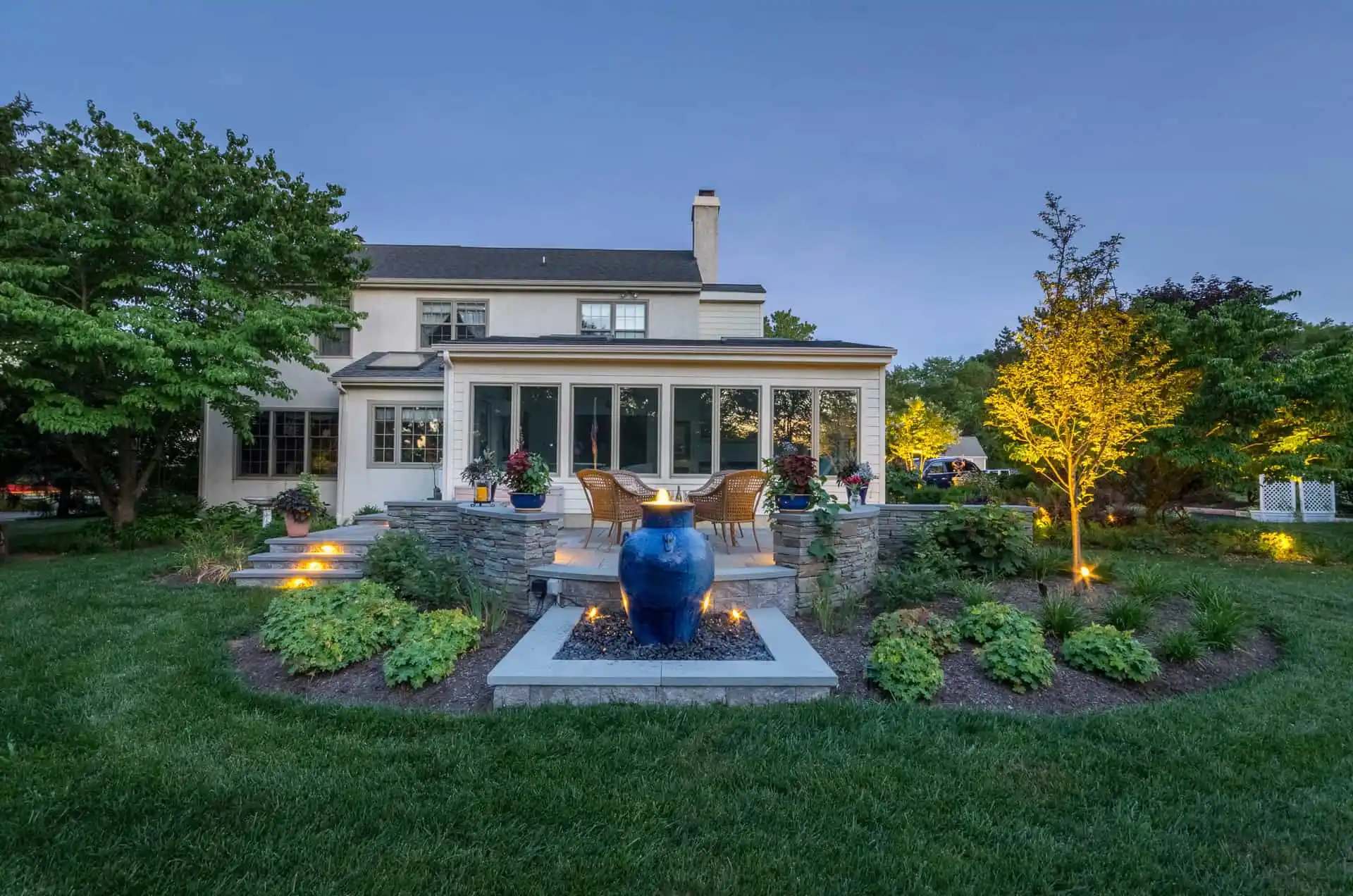 A well-lit backyard garden with a blue ceramic fountain centerpiece, surrounded by landscaped plants, a stone patio, outdoor seating, and a two-story house at dusk.