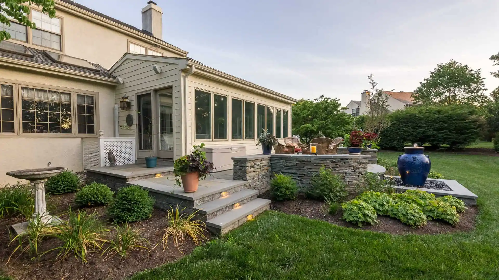 A suburban home with a sunroom extension offers cozy outdoor living in Delaware County, PA, featuring a seating area surrounded by stone walls. The garden's lush greenery, bushes, birdbath, and large blue ceramic vase create an inviting and serene backyard oasis.