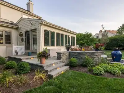 A suburban home with a sunroom extension offers cozy outdoor living in Delaware County, PA, featuring a seating area surrounded by stone walls. The garden's lush greenery, bushes, birdbath, and large blue ceramic vase create an inviting and serene backyard oasis.