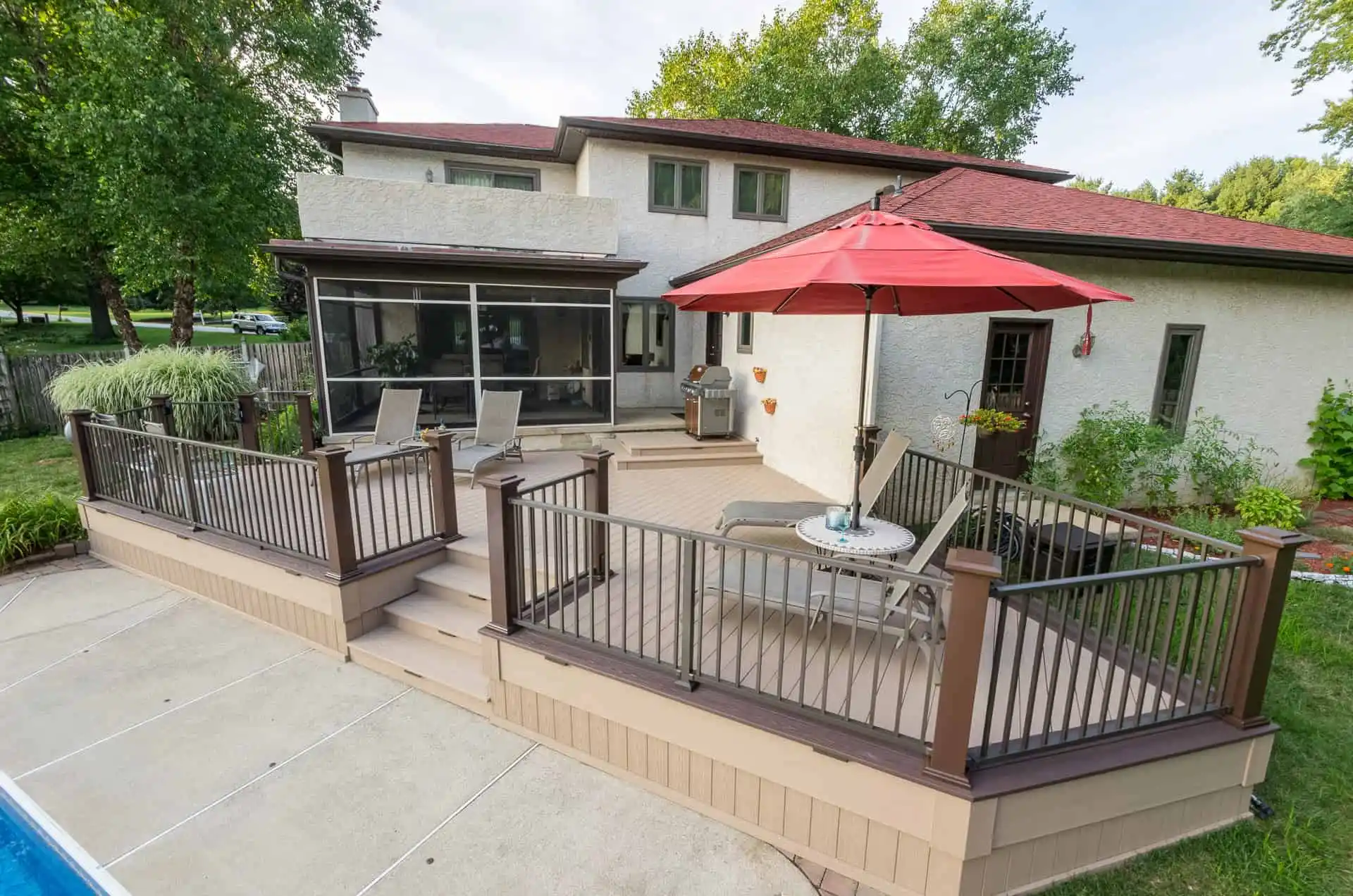 A backyard deck with brown railings, lounge chairs, a table with a red umbrella, and stairs leading to a pool. The deck is attached to a two-story house surrounded by green trees and grass.