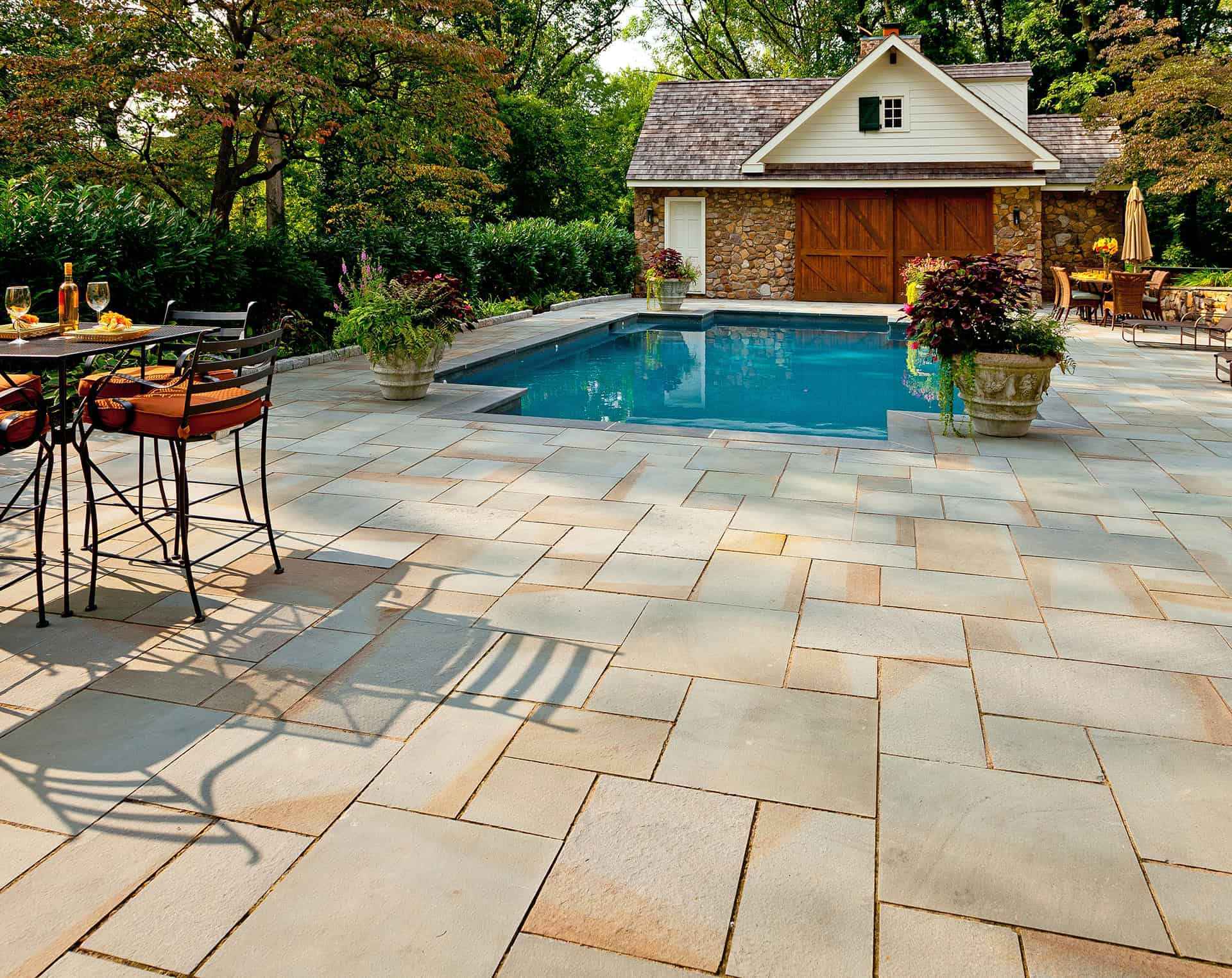 A backyard scene with a swimming pool, stone patio, potted plants, a table with chairs, and a small stone pool house with wooden doors, surrounded by trees and greenery.