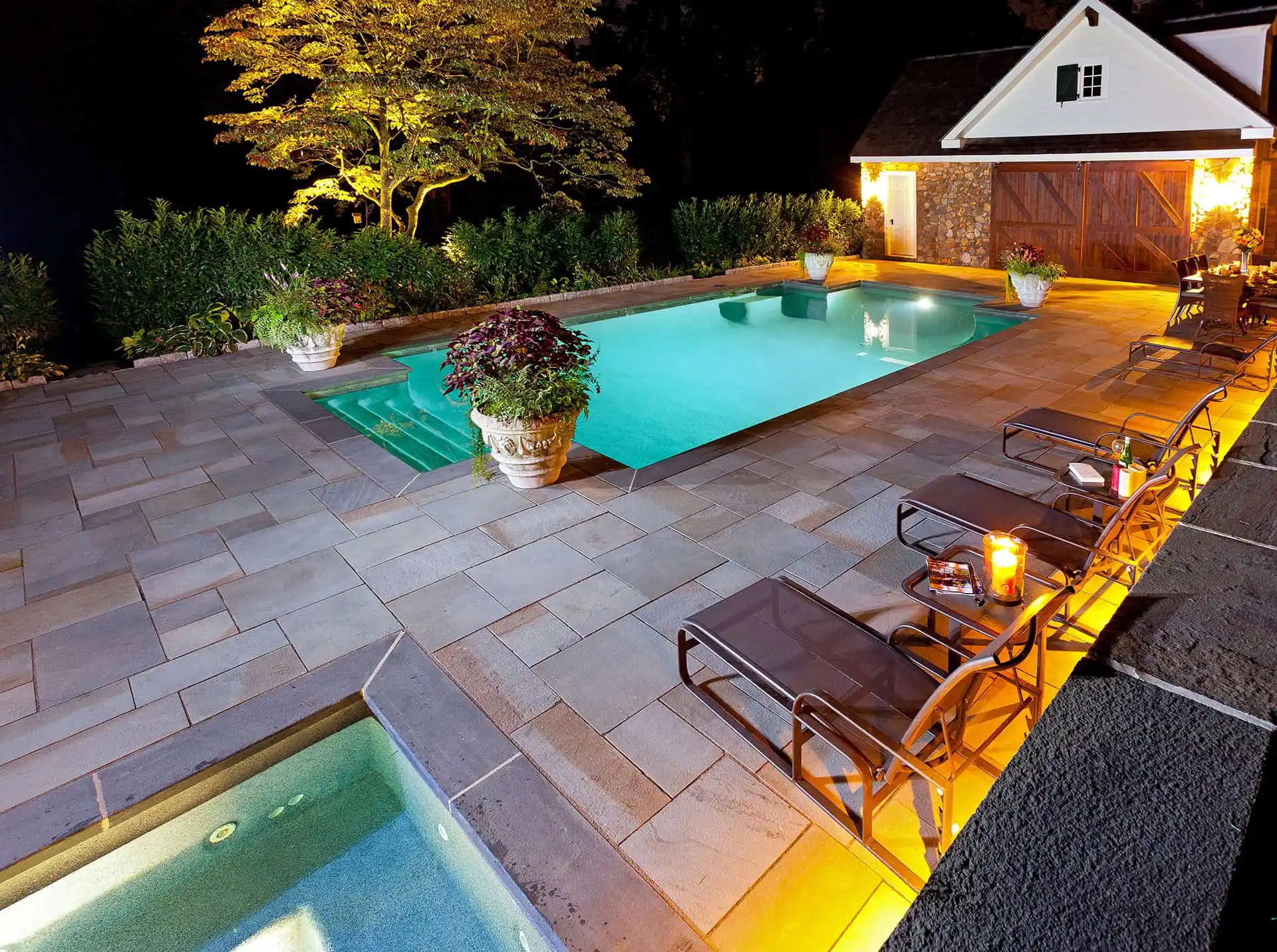 A well-lit backyard with a rectangular swimming pool, lounge chairs, a hot tub in the foreground, large potted plants, and a house with open wooden doors in the background at night.