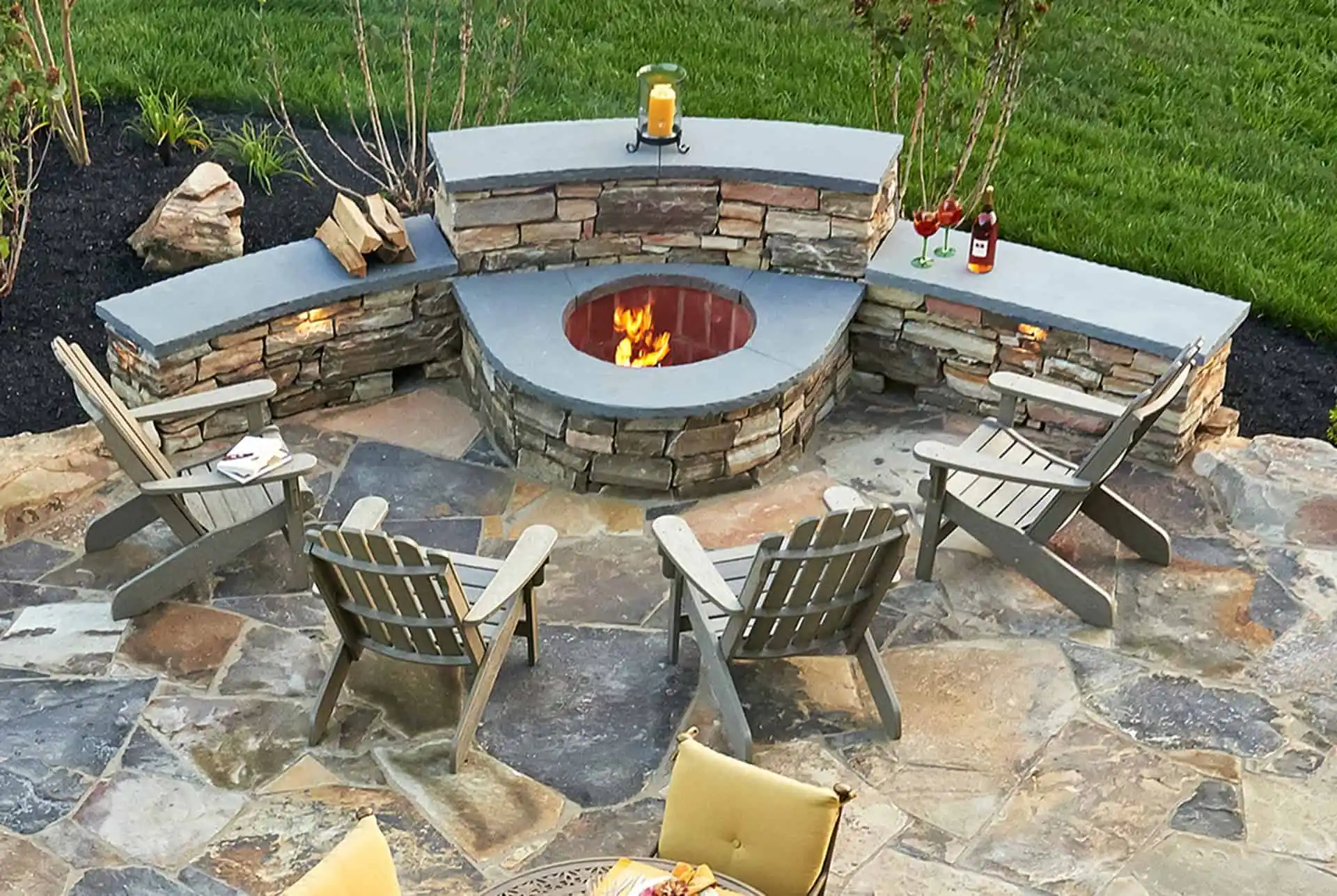 Outdoor patio with a stone fire pit surrounded by four wooden chairs and curved stone benches. Drinks and a candle are on the benches, and the fire pit is lit. The patio is tiled with stone and opens to green grass.