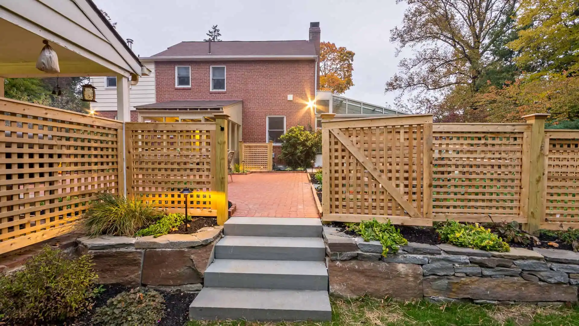 Two-story brick house with a wooden lattice fence enclosing a red brick pathway. Stone steps lead to the gate, surrounded by landscaped greenery from top-notch landscaping services in Delaware County, PA. A tall tree stands to the right, gently lit by outdoor lights.