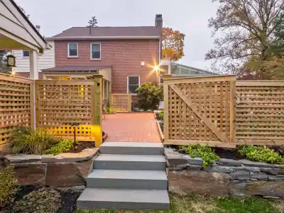 Two-story brick house with a wooden lattice fence enclosing a red brick pathway. Stone steps lead to the gate, surrounded by landscaped greenery from top-notch landscaping services in Delaware County, PA. A tall tree stands to the right, gently lit by outdoor lights.