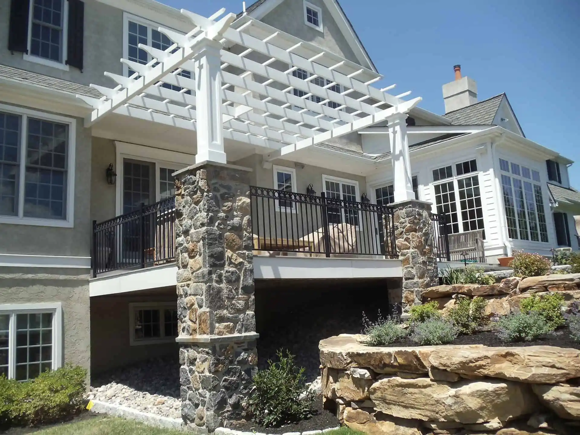 A two-story house with a raised patio featuring stone pillars, black railings, and a white pergola. The patio overlooks a landscaped yard with stone retaining walls and green plants.