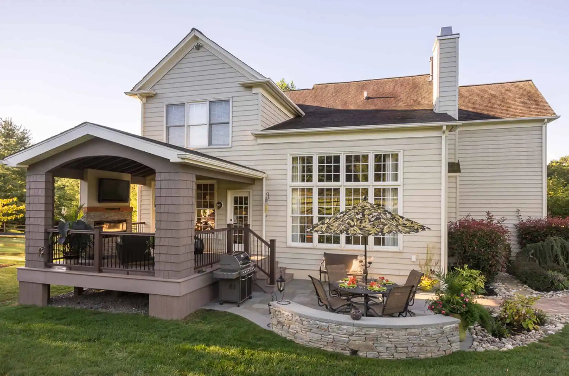 A beige two-story house with large windows, a covered porch with seating and a TV, a patio with an umbrella, table and chairs, a grill, landscaped plants, and a lush green lawn in the foreground.