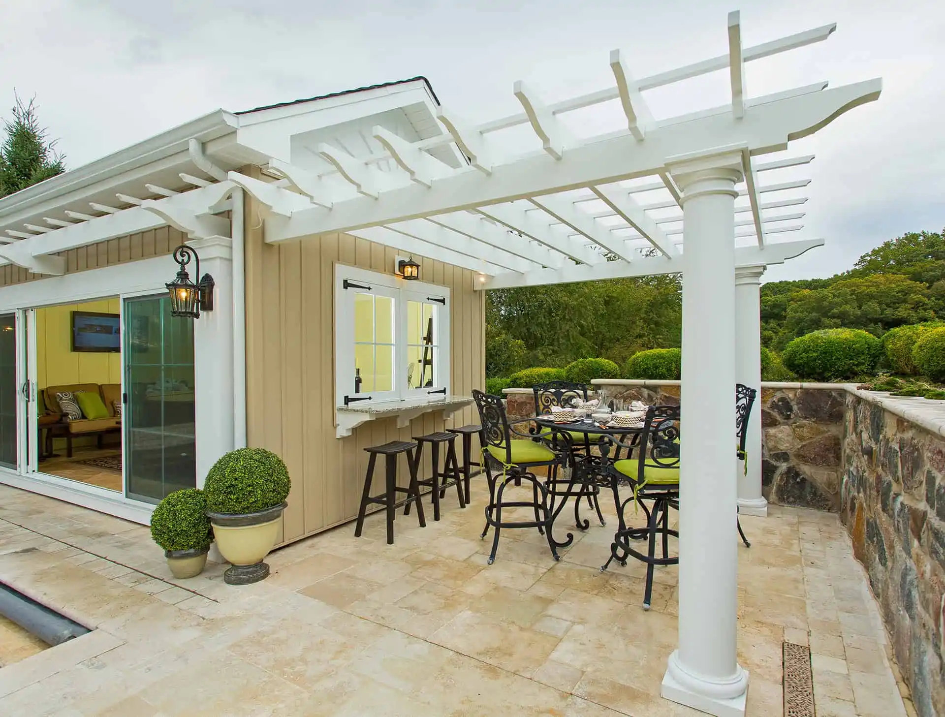 A stylish outdoor patio with a white pergola, tall chairs around a dining table, and potted topiary plants. A sliding glass door leads inside, and a stone wall borders the space, overlooking a green landscape.