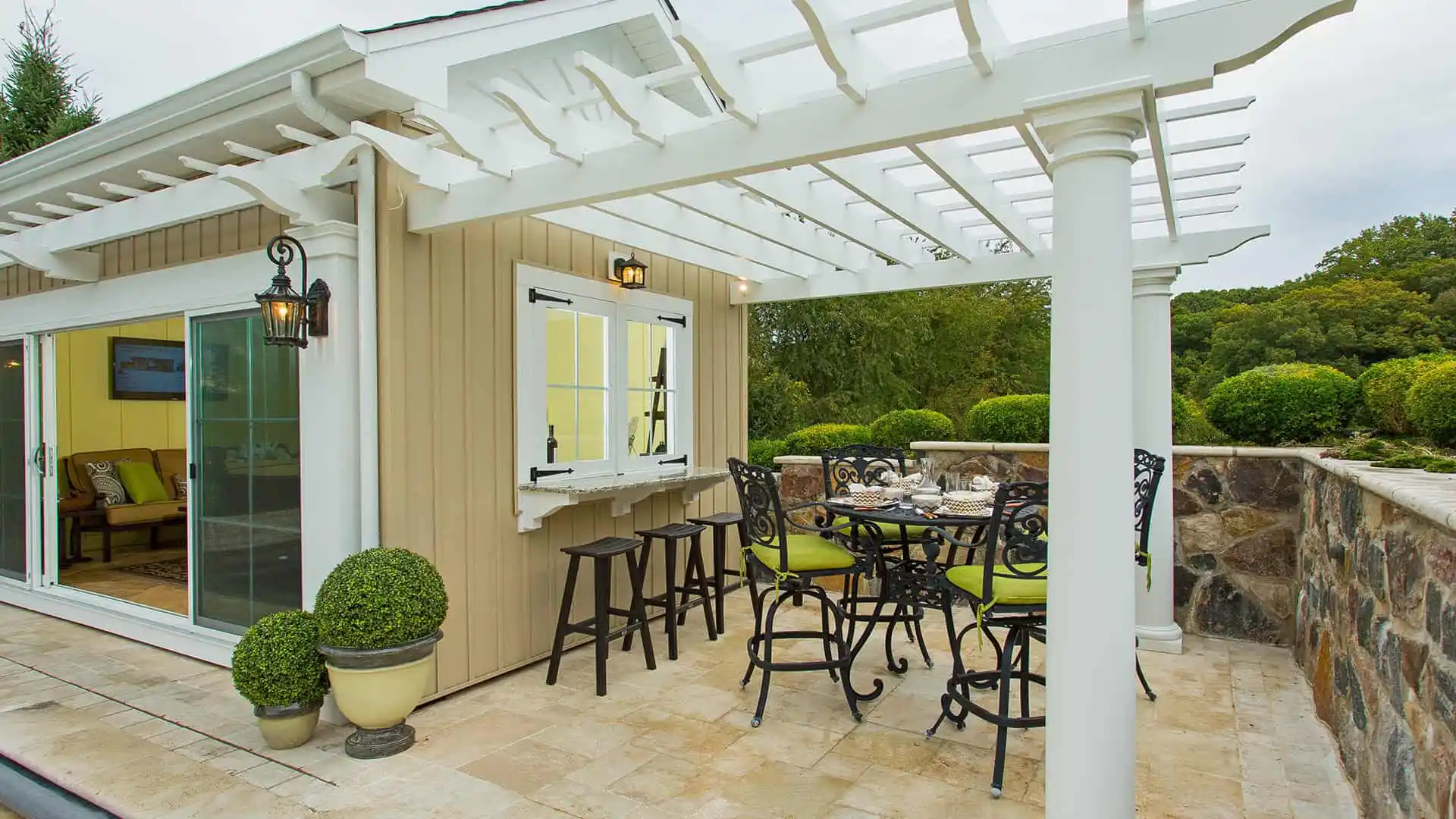 A cozy outdoor patio with a pergola attached to a house showcases the charm of outdoor living in Delaware County, PA. It features a round dining table with wrought-iron chairs adorned with green cushions. Potted plants enhance the space, while trees and a large window counter complete the inviting scene.