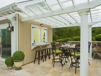 A cozy outdoor patio with a pergola attached to a house showcases the charm of outdoor living in Delaware County, PA. It features a round dining table with wrought-iron chairs adorned with green cushions. Potted plants enhance the space, while trees and a large window counter complete the inviting scene.