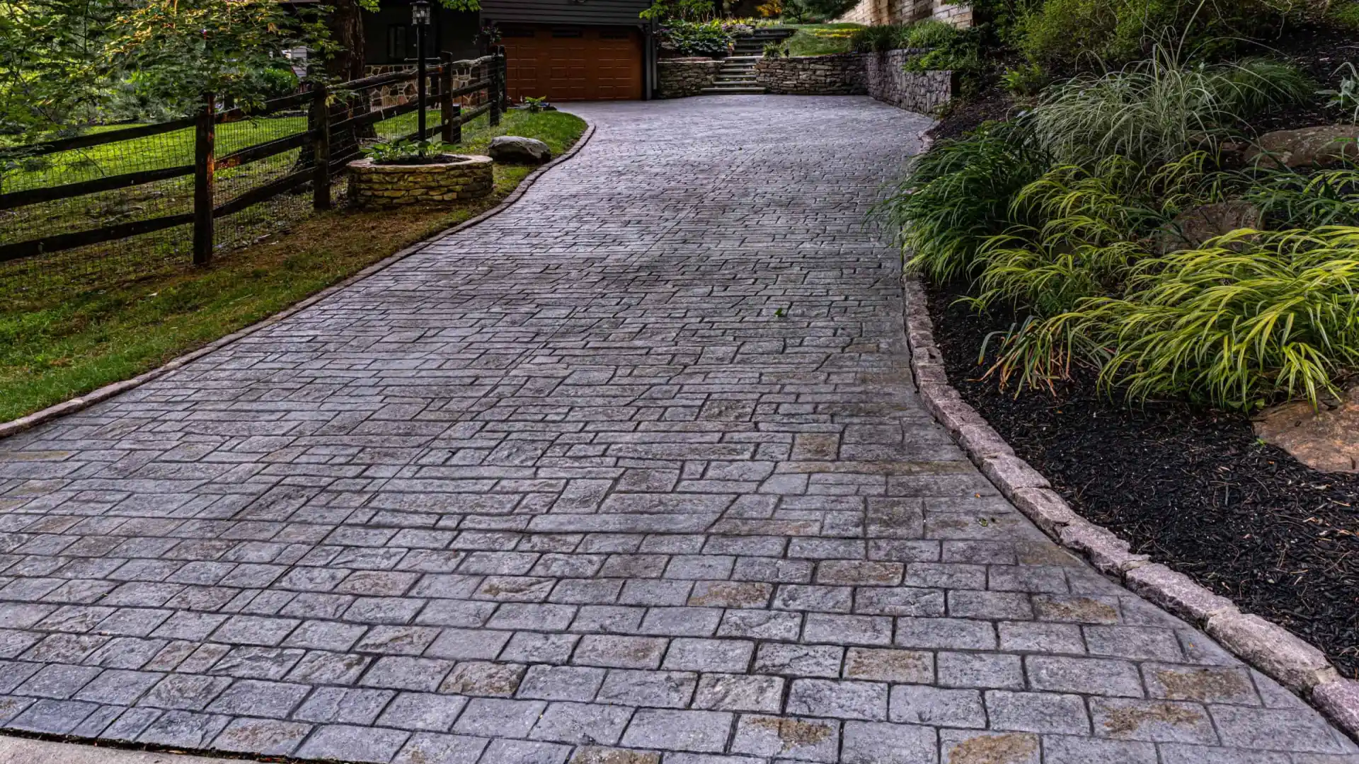 A long, winding driveway of gray interlocking bricks leads to a wooden garage, showcasing exquisite hardscaping services Delaware County PA. Lush greenery lines both sides, with a wooden fence on the left and stone retaining walls on the right, beautifully bordered by vibrant plants.