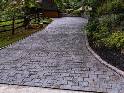 A long, winding driveway of gray interlocking bricks leads to a wooden garage, showcasing exquisite hardscaping services Delaware County PA. Lush greenery lines both sides, with a wooden fence on the left and stone retaining walls on the right, beautifully bordered by vibrant plants.
