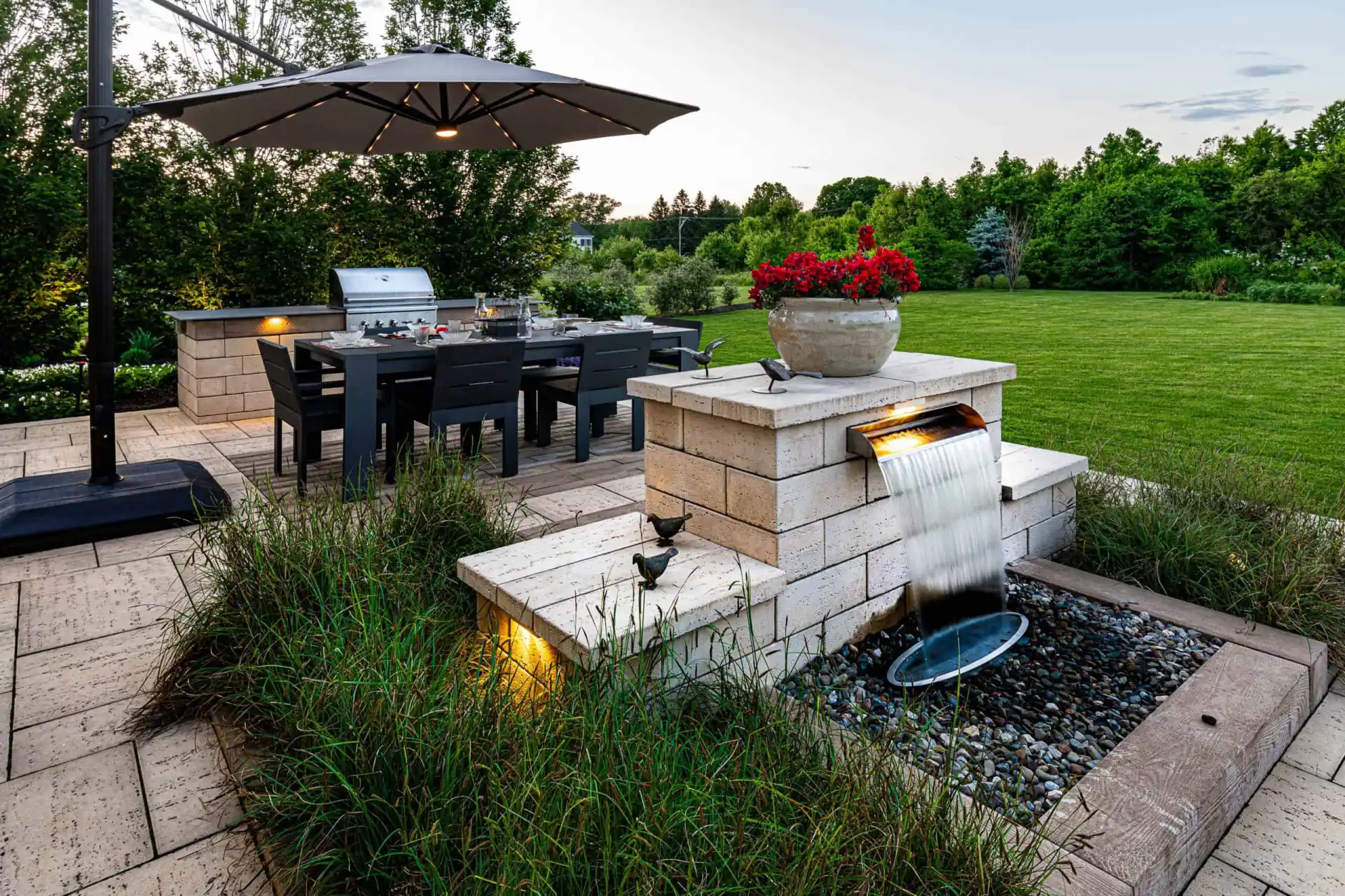 A backyard patio with a dining table and chairs sits on a tiled surface, showcasing expert hardscaping services in Delaware County, PA. A small water feature with a cascade is nearby, surrounded by grassy plants. An umbrella provides shade, overlooking a lawn and tree-lined background.