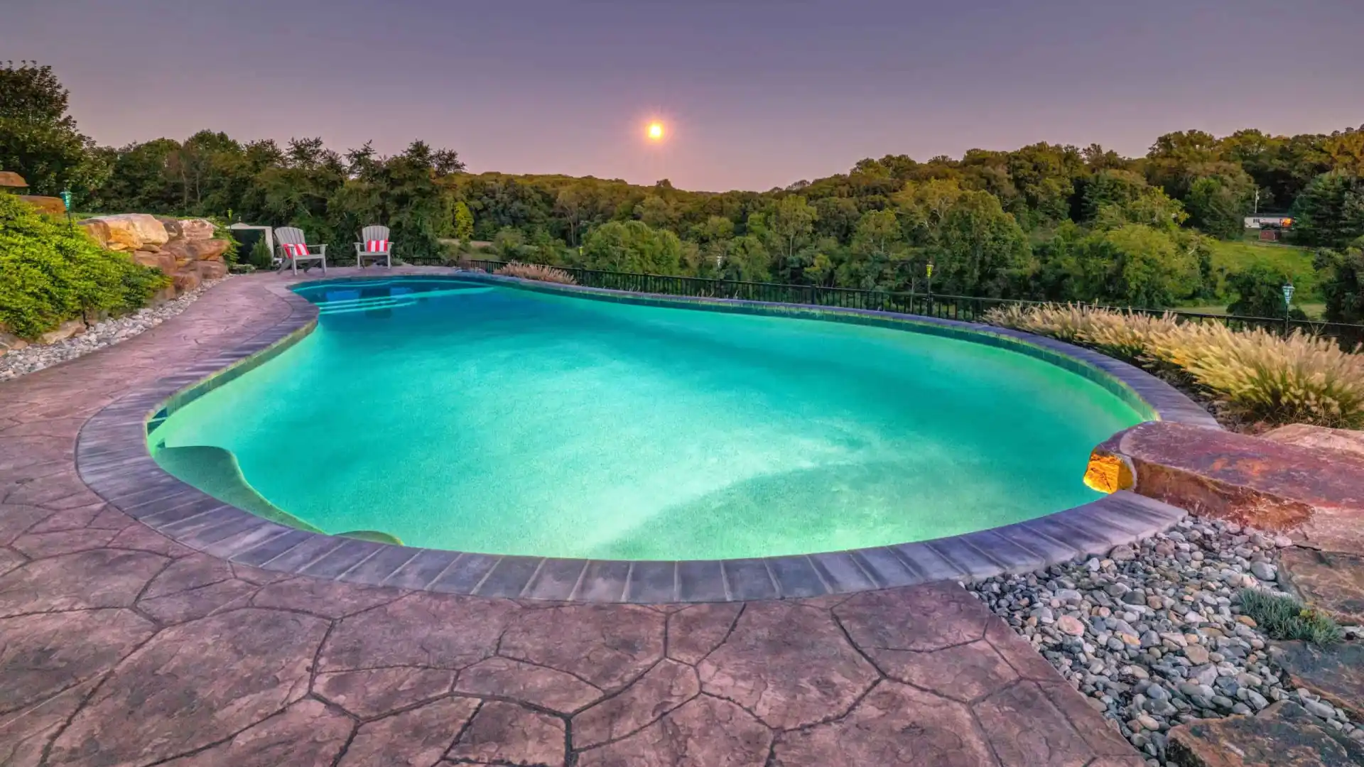 A tranquil swimming pool glows under a serene sky at sunset, surrounded by lush greenery thanks to expert landscaping services in Delaware County, PA. Two chairs on a stone patio at the far end create a peaceful and inviting outdoor living atmosphere.