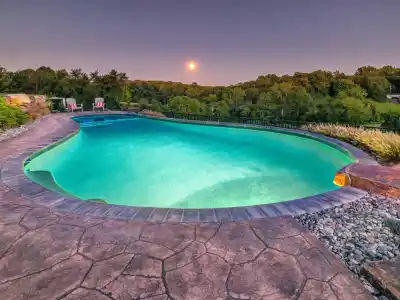 A tranquil swimming pool glows under a serene sky at sunset, surrounded by lush greenery thanks to expert landscaping services in Delaware County, PA. Two chairs on a stone patio at the far end create a peaceful and inviting outdoor living atmosphere.