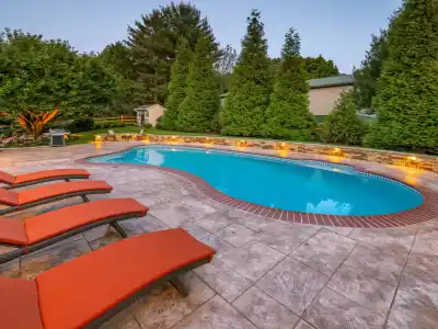 A backyard scene in Delaware County, PA, showcases a kidney-shaped swimming pool nestled within a beautifully paved patio. Four red-orange cushioned lounge chairs invite relaxation, while tall evergreens stand proudly in the background. Expert hardscaping services enhance the cozy ambiance with small lights illuminating the area.