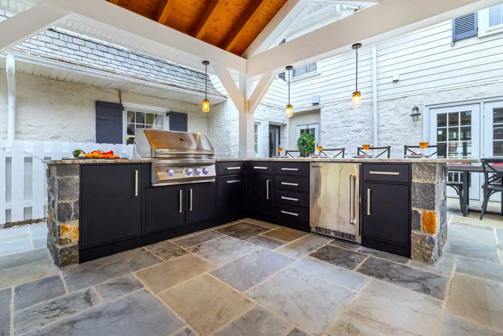 Outdoor living in Delaware County, PA is elevated with this kitchen featuring stone flooring and black cabinetry. It includes a built-in grill, sink, and countertop under a wooden pergola. A table set near the white house and picket fence enjoys gentle illumination from hanging lights.
