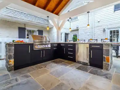 Outdoor living in Delaware County, PA is elevated with this kitchen featuring stone flooring and black cabinetry. It includes a built-in grill, sink, and countertop under a wooden pergola. A table set near the white house and picket fence enjoys gentle illumination from hanging lights.