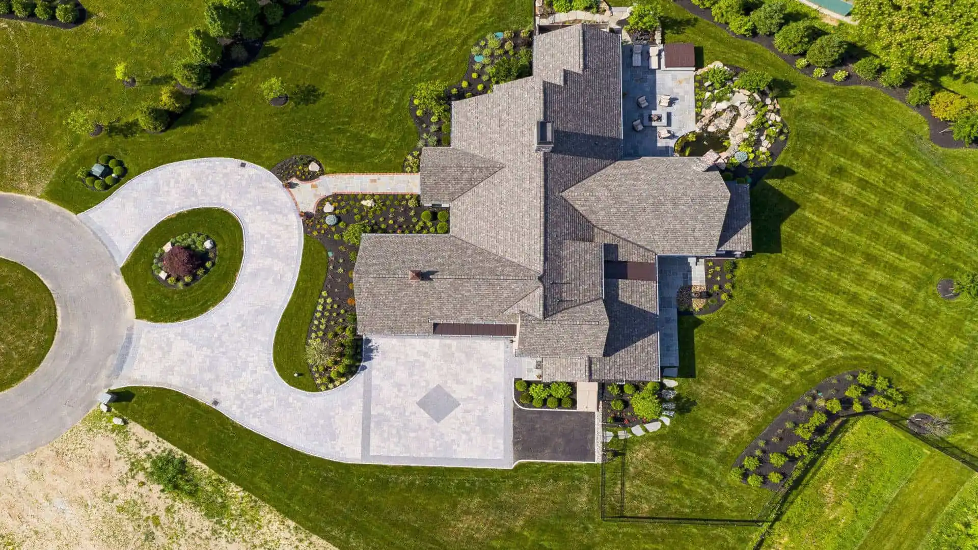 Aerial view of a large house with a grey roof, nestled amid expertly landscaped lawn by renowned landscaping services in Delaware County, PA. A circular driveway leads to the entrance, complemented by a backyard patio area. Trees and shrubs enhance this outdoor living haven.