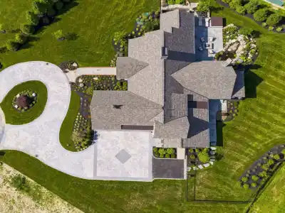 Aerial view of a large house with a grey roof, nestled amid expertly landscaped lawn by renowned landscaping services in Delaware County, PA. A circular driveway leads to the entrance, complemented by a backyard patio area. Trees and shrubs enhance this outdoor living haven.