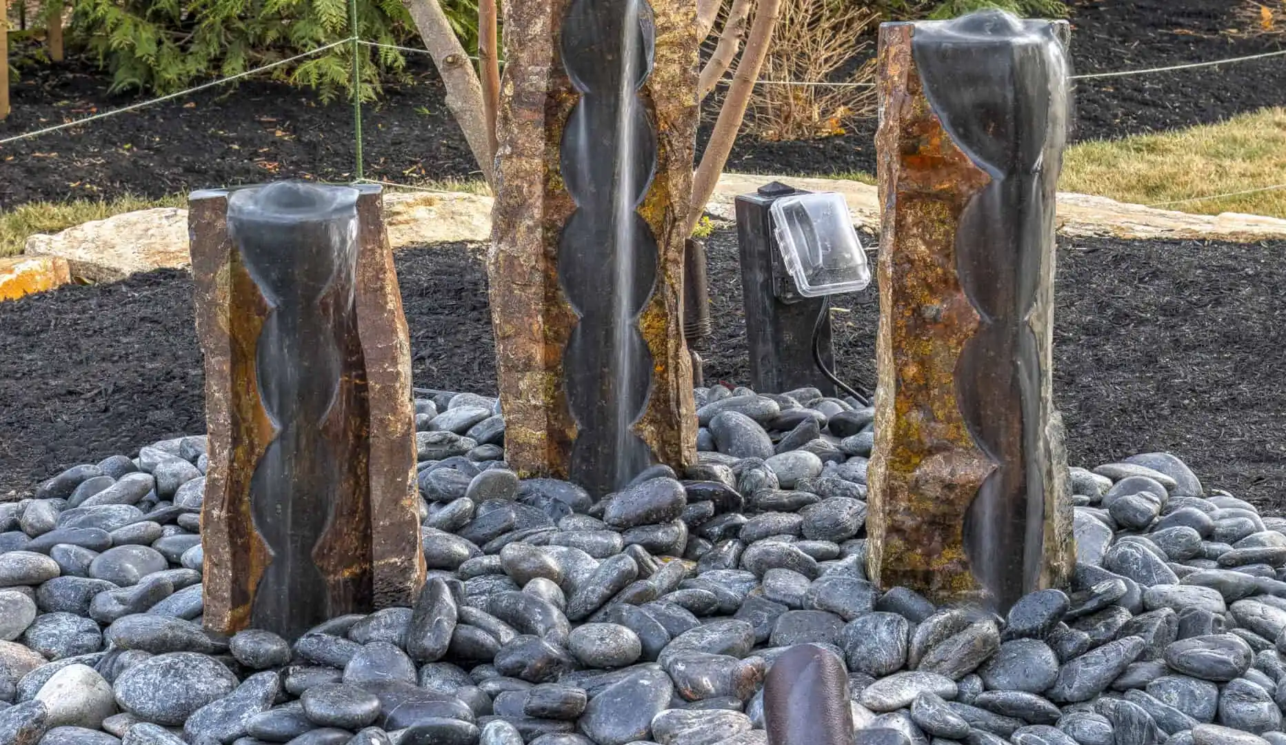 Three vertical stone fountains, encircled by smooth black pebbles, grace a landscaped garden. A tree with yellowish leaves stands behind while outdoor lighting softly illuminates the area. This serene setting is beautifully crafted by premier landscaping services in Delaware County, PA.