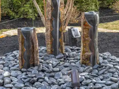 Three vertical stone fountains, encircled by smooth black pebbles, grace a landscaped garden. A tree with yellowish leaves stands behind while outdoor lighting softly illuminates the area. This serene setting is beautifully crafted by premier landscaping services in Delaware County, PA.